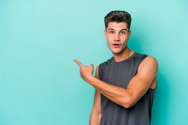 Mavi arka planda izole edilmiş genç beyaz adam gülümsüyor ve boş alanda bir şeyler gösteriyor..