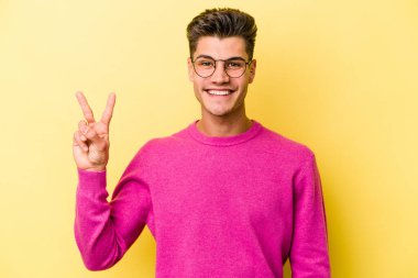 Sarı arka planda izole edilmiş genç beyaz adam zafer işareti gösteriyor ve gülümsüyor..
