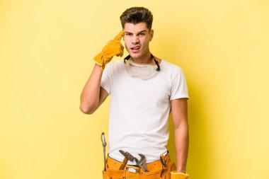 Sarı arka planda izole edilmiş genç bir elektrikçi işaret parmağıyla hayal kırıklığı gösteriyor..