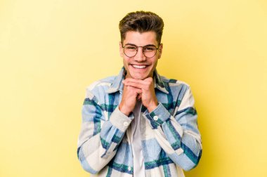 Sarı arka planda izole edilmiş genç beyaz adam ellerini çenesinin altında tutuyor ve mutlu bir şekilde yan tarafa bakıyor..