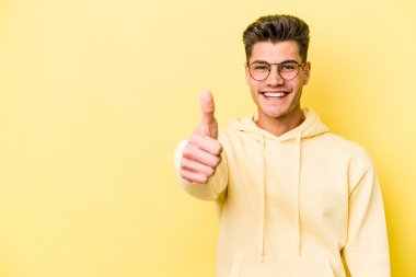 Sarı arka planda izole edilmiş genç beyaz adam gülümsüyor ve başparmağını kaldırıyor.