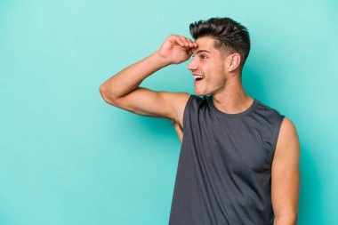 Kafkasyalı genç adam mavi arka planda izole edilmiş, uzaklara bakıyor ve elini alnına koyuyor..