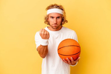 Genç basketbolcu beyaz adam sarı arka planda izole edilmiş kameraya yumruk gösteriyor, agresif yüz ifadesi var..