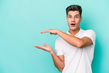 Mavi arka planda izole genç beyaz adam şok ve hayret içinde elleri arasında bir kopya boşluk tutuyor.