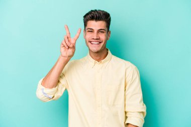 Mavi arka planda izole edilmiş genç beyaz adam iki numarayı parmaklarıyla gösteriyor..