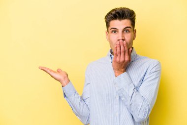 Sarı arka planda izole edilmiş genç beyaz adam, avuç içi fotokopi alanını elinde tutmaktan etkilendi..