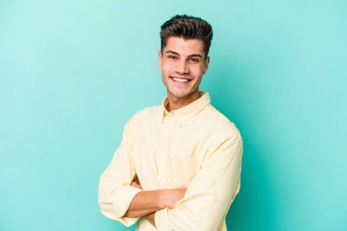 Mavi arka planda kendini güvende hisseden, kararlılıkla kollarını kavuşturan genç beyaz bir adam..