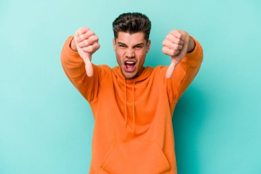 Mavi arka planda izole edilmiş genç beyaz bir adam baş parmağını aşağı gösteriyor ve hoşlanmadığını ifade ediyor..