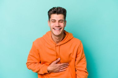 Mavi arka planda izole olmuş genç beyaz adam yüksek sesle gülüyor ve elini göğsüne koyuyor..