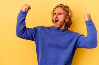 Sarı arka planda izole edilmiş genç beyaz adam zaferden sonra yumruğunu kaldırıyor, kazanan konsept..