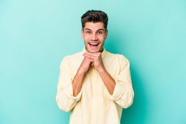 Kafkasyalı genç adam mavi arka planda izole edilmiş şans için dua ediyor, şaşırmış ve ağzını açıp önüne bakıyor..