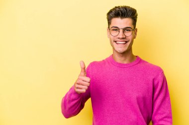 Sarı arka planda izole edilmiş genç beyaz adam gülümsüyor ve başparmağını kaldırıyor.
