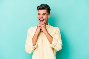 Mavi arka planda izole edilmiş genç beyaz adam ellerini çenesinin altında tutuyor, mutlu bir şekilde yana bakıyor..