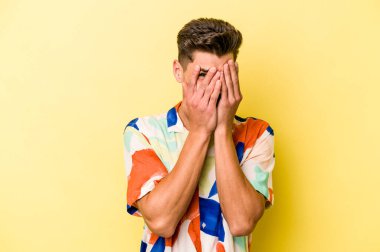 Sarı arka planda izole edilmiş genç beyaz adam kameraya parmaklarıyla göz kırpıyor, yüzünü kapatmaktan utanıyor..