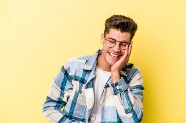 Sarı arka planda izole edilmiş genç beyaz adam mutlu bir şekilde gülüyor ve ellerini karnının üstünde tutarak eğleniyor..