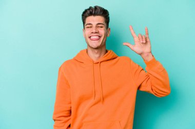 Mavi arka planda izole edilmiş genç beyaz adam parmaklarıyla kaya hareketi gösteriyor.