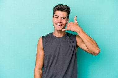 Mavi arka planda izole edilmiş genç beyaz adam parmaklarıyla bir cep telefonu hareketi gösteriyor..