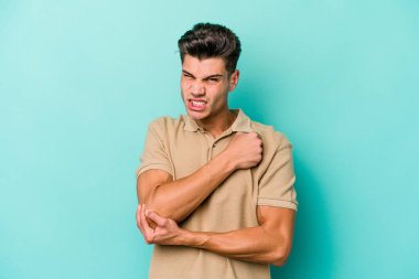 Kafkasyalı genç adam mavi arka planda izole edilmiş dirsek masajı yapıyor, kötü bir hareketin ardından acı çekiyor..