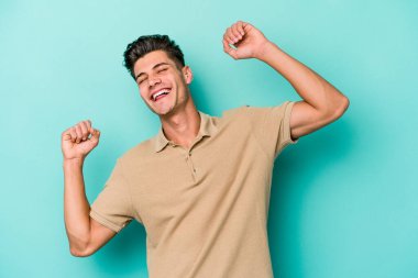 Mavi arka planda izole genç beyaz adam özel bir günü kutluyor, atlıyor ve enerjiyle kollarını kaldırıyor..