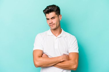 Kafkasyalı genç adam mavi arka planda izole edilmiş hoşnutsuzca kaşlarını çatmış, kollarını kavuşturmuş..