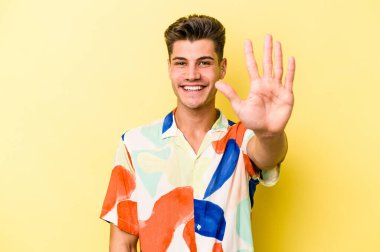 Sarı arka planda izole edilmiş genç beyaz adam gülümseyerek beş numarayı parmaklarıyla gösteriyor..