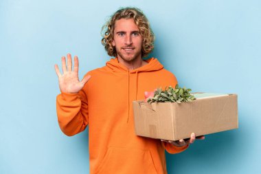 Kafkasyalı genç adam mavi arka planda izole edilmiş şeylerle dolu bir kutuyu kaldırırken hareket ediyor. Beş numarayı parmaklarıyla gülümsüyor..