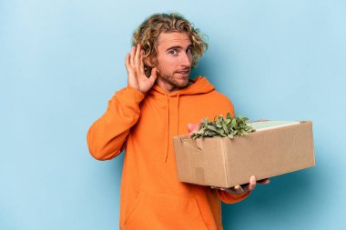 Genç beyaz adam, mavi arka planda izole edilmiş şeylerle dolu bir kutuyu alıp bir dedikoduyu dinlerken hamle yapıyor..