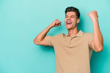 Mavi arka planda izole edilmiş genç beyaz bir adam zaferden sonra yumruğunu kaldırıyor, kazanan konsept..