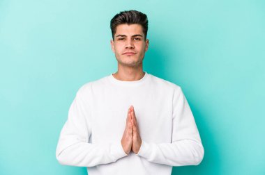 Kafkasyalı genç adam mavi arka planda izole edilmiş dua ediyor, bağlılık gösteriyor, dindar biri ilahi ilham arıyor..