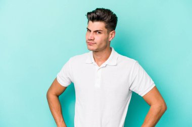 Kafkasyalı genç adam mavi arka planda izole, kafası karışmış, şüpheli ve güvensiz hissediyor..