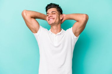 Kafkasyalı genç adam mavi arka planda izole edilmiş. Kendine güveniyor, elleri başının arkasında..