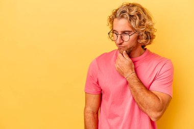 Sarı arka planda izole edilmiş genç beyaz adam şüpheli ve şüpheci bir ifadeyle yana bakıyor..
