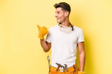 Genç elektrikçi beyaz adam sarı arka plan noktalarında izole edilmiş başparmak uzakta, gülüyor ve kaygısız.