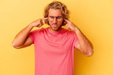 Sarı arka planda izole edilmiş genç beyaz bir adam parmaklarıyla kulaklarını kapatıyor, gürültülü bir ortam yüzünden stresli ve çaresiz..