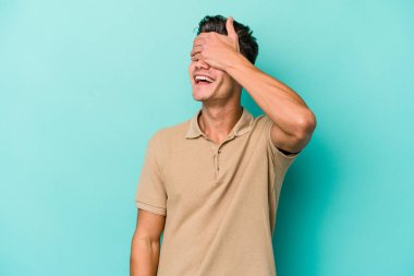 Mavi arka planda soyutlanmış genç beyaz bir adam elleriyle gözlerini kapatıyor, geniş bir gülümsemeyle bir sürpriz bekliyor..