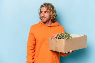 Kafkasyalı genç bir adam mavi arka planda izole edilmiş eşyalarla dolu bir kutuyu alırken gülümsüyor, neşeli ve keyifli görünüyor..