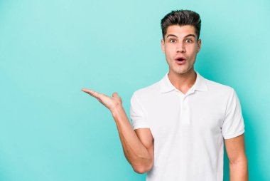 Mavi arka planda izole edilmiş genç beyaz adam, avuç içi fotokopi alanını elinde tutmaktan etkilenmiş..