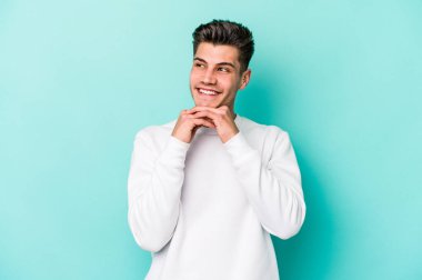 Mavi arka planda izole edilmiş genç beyaz adam ellerini çenesinin altında tutuyor, mutlu bir şekilde yana bakıyor..