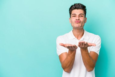Kafkasyalı genç adam mavi arka planda izole edilmiş dudaklar kıvrılıyor ve hava öpücüğü göndermek için avuç içleri tutuyor..
