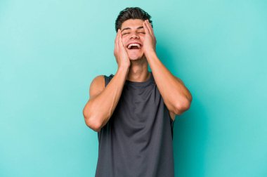 Mavi arka planda izole edilmiş genç beyaz adam ellerini başının üstünde tutarak neşe içinde gülüyor. Mutluluk kavramı.