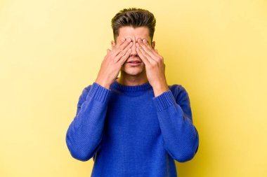 Sarı arka planda izole edilmiş genç beyaz adam elleriyle gözlerini kapatmaktan korkuyor..