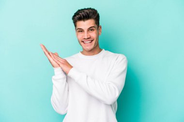 Kafkasyalı genç adam mavi arka planda izole edilmiş enerjik ve rahat hissediyor, ellerini ovuşturuyor kendinden emin.