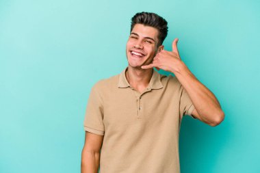 Mavi arka planda izole edilmiş genç beyaz adam parmaklarıyla bir cep telefonu hareketi gösteriyor..