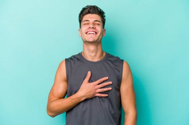 Mavi arka planda izole olmuş genç beyaz adam yüksek sesle gülüyor ve elini göğsüne koyuyor..