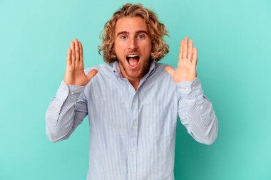 Mavi arka planda izole edilmiş genç beyaz adam hoş bir sürpriz alıyor, heyecanlı ve el kaldırıyor..