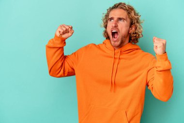 Mavi arka planda izole edilmiş genç beyaz bir adam zaferden sonra yumruğunu kaldırıyor, kazanan konsept..