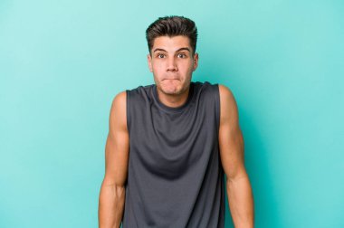 Mavi arka planda izole edilmiş genç beyaz adam omuzlarını silkeliyor ve gözleri açık..