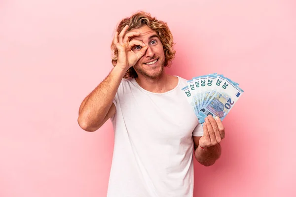 Young caucasian man holding banknotes isolated on pink background excited keeping ok gesture on eye.