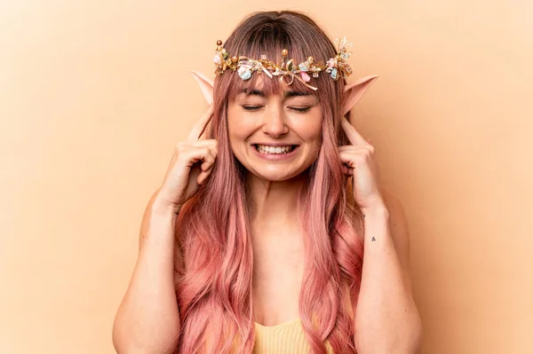 Young elf woman with pink hair isolated on beige background covering ears with hands.