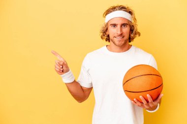 Genç basketbolcu beyaz adam sarı arka planda izole edilmiş gülümsüyor ve boş alanda bir şeyler gösteriyor..
